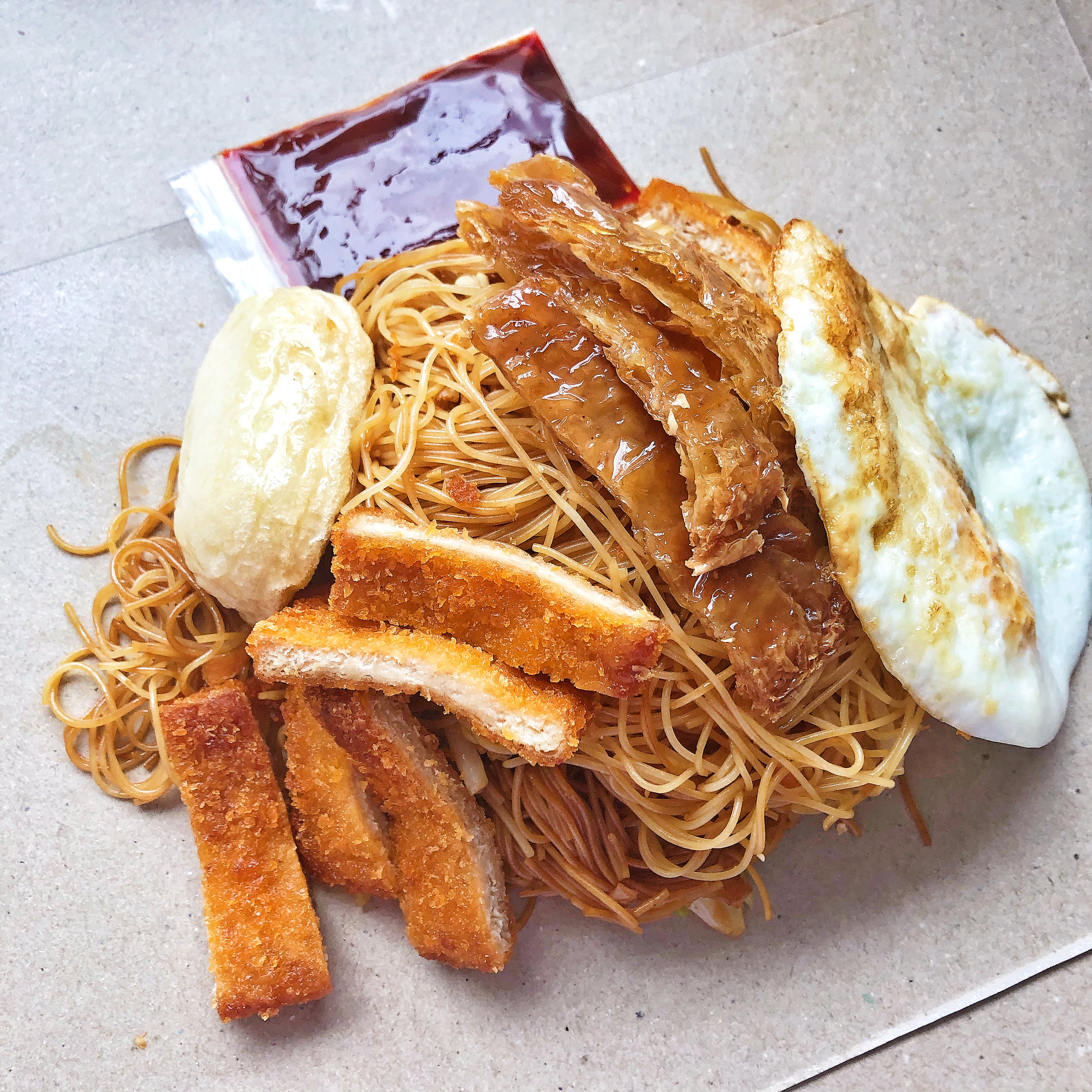 Thin rice noodles in soy sauce with fried fish cake, fish sticks and fried egg with a packet of chilli sauce